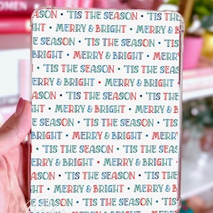 以下が含まれることがあります： 透明な携帯電話ケース。白地に赤、青、緑で「'Tis the Season」と「Merry & Bright」のフレーズが繰り返されるパターン。ケースは手で持たれています。