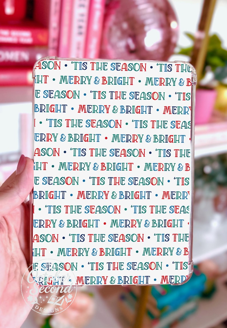 以下が含まれることがあります： 透明な携帯電話ケース。白地に赤、青、緑で「'Tis the Season」と「Merry & Bright」のフレーズが繰り返されるパターン。ケースは手で持たれています。