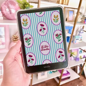 Peut inclure: Une liseuse électronique noire avec une coque de protection transparente. L'écran affiche un motif de rayures verticales bleu sarcelle avec des cadres ovales contenant diverses illustrations florales et le texte KB's Kindle en rose foncé.