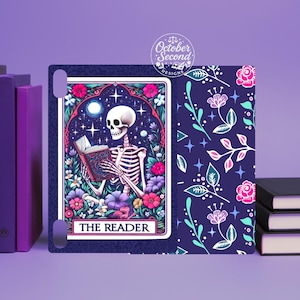 May include: A tarot card featuring a skeleton reading a book, surrounded by flowers and a crescent moon. The card is framed in a decorative border with the words "THE READER" at the bottom. The background is a dark blue with floral patterns.