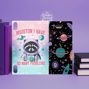May include: Two decorative book covers. One features a raccoon astronaut with the text "Houston I Have So Many Problems" on a pastel background. The other has a black background with colorful planets and stars.
