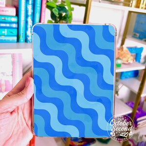 May include: A clear tablet case featuring a wavy pattern in shades of blue. The design is composed of alternating light and dark blue curved lines. The case has rounded corners and a logo that reads "October Second Designs."