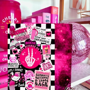 May include: A pink and black decorative item with a checkerboard pattern and various stickers, including text that reads "Bookish Bitches Go To Heaven." A pink disco ball and books are in the background.