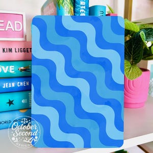 May include: A blue notebook with a wavy pattern in shades of blue. The notebook is on a shelf with books and a pink pot with a green plant. The text "October Second Designs" is visible.