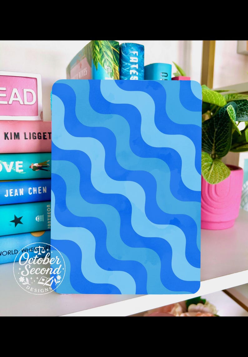 May include: A blue notebook with a wavy pattern in shades of blue. The notebook is on a shelf with books and a pink pot with a green plant. The text "October Second Designs" is visible.