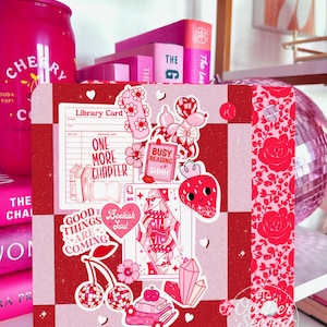 May include: A decorative pink and red bookish-themed display. The display features a checkerboard pattern with stickers of a library card, a queen of hearts, and a strawberry. The background includes pink books and a cherry soda can.