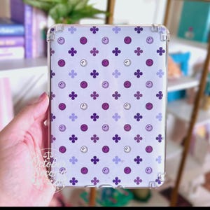 May include: Clear protective case for a tablet with a white background. The case features a pattern of purple smiley faces and four-petal flowers. The corners of the case are reinforced for added protection. The text "October Second Designs" is visible.
