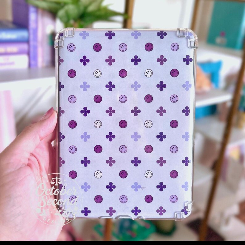 May include: Clear protective case for a tablet with a white background. The case features a pattern of purple smiley faces and four-petal flowers. The corners of the case are reinforced for added protection. The text "October Second Designs" is visible.