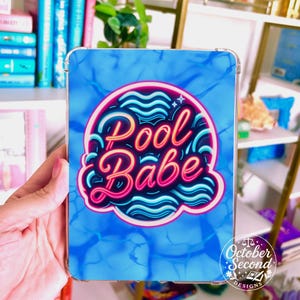 May include: A clear phone case featuring a blue water pattern background. The case has a neon pink and blue design that says "Pool Babe". The October Second Designs logo is in the bottom right corner.