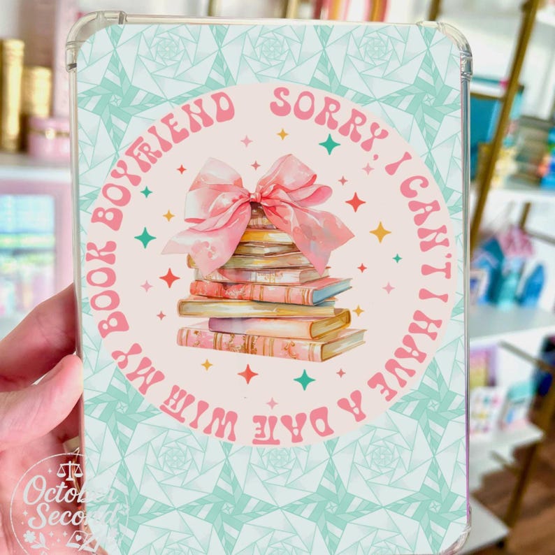 A hand holding a book cover with a playful design featuring a stack of books with a pink bow and stars around it.
