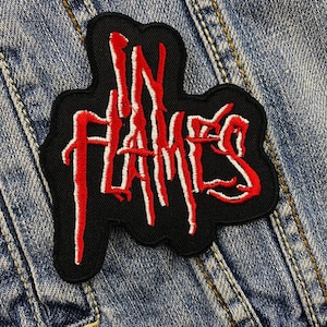 Könnte beinhalten: Schwarzer, gestickter Aufnäher mit den roten und weißen Worten "IN FLAMES". Der Aufnäher hat einen schwarzen Rand und befindet sich auf einem Jeansstoff-Hintergrund.
