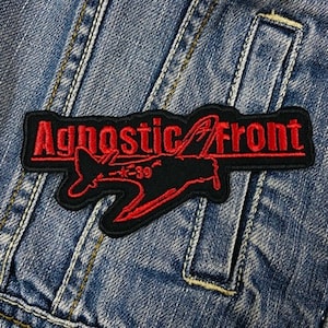Puede incluir: Un parche bordado en negro y rojo con el texto "Agnostic Front" y un diseño estilizado de avión. El parche está cosido sobre una chaqueta vaquera azul, mostrando la textura de la tela y los detalles de las costuras.