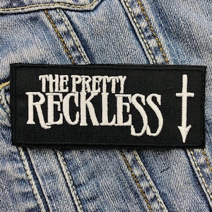Op de afbeelding: Een zwart rechthoekig embleem met de witte geborduurde tekst "THE PRETTY RECKLESS" en een wit kruis met een pijl naar beneden. Het embleem is geplaatst op een blauwe denim achtergrond.