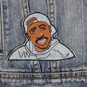Puede incluir: Parche bordado con un retrato de dibujos animados de un hombre con gorra y sudadera con capucha. El parche tiene un esquema de color azul claro, marrón y morado, con contornos negros. Cosido en una chaqueta vaquera.