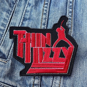 Puede incluir: Parche bordado con las palabras "Thin Lizzy" en rojo, contorneadas en blanco, sobre un fondo negro. El parche incluye una silueta roja de un guitarrista. El parche está cosido a una chaqueta vaquera.