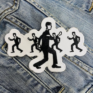 Op de afbeelding: Geborduurde patch met een zwart-wit ontwerp van figuren in pakken. De patch heeft een witte rand en is op een spijkerjasje geplaatst. De figuren zijn in beweging, met uitgestrekte armen en gebogen benen.