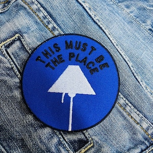 May include: A round, blue patch with the text "THIS MUST BE THE PLACE" in black lettering. A white lamp graphic is centered on the patch. The patch is sewn onto a denim jacket.