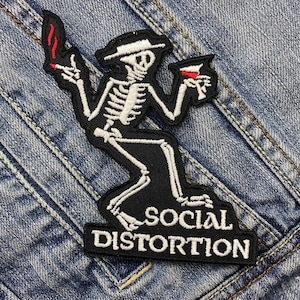 May include: Black and white embroidered patch featuring a skeleton figure holding a lit cigarette and a drink. The skeleton wears a hat and is set against a black background. The words "SOCIAL DISTORTION" are embroidered below the figure.