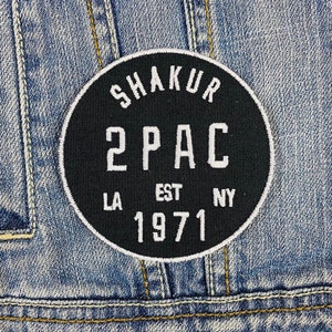 May include: A black circular patch with white text reading "SHAKUR 2 PAC EST 1971 LA NY" is sewn onto a blue denim jacket. The patch has a white border and is centered on the jacket's back.