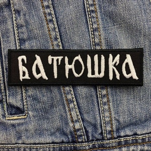May include: Black rectangular patch with the white word "БАТЮШКА" embroidered on it. The patch is sewn onto a blue denim jacket. The text is in a stylized font.