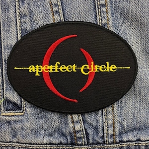 Op de afbeelding: Zwarte ovale patch met de tekst "aperfect circle" in geel, met een rood halvemaanvormig ontwerp. De patch is op een spijkerjasje.