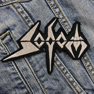 May include: Embroidered patch featuring the word "SODOM" in a stylized, angular font. The patch has a black border with the text in light gray. The patch is set against a blue denim background.