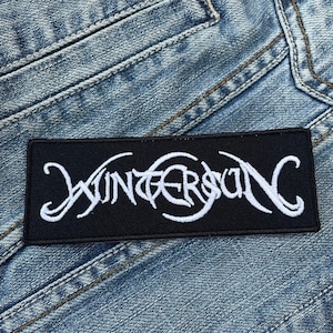 May include: A black, rectangular patch featuring the white embroidered word "WINTERSUN" in a stylized font. The patch is placed on a light blue denim fabric, with visible stitching and seams.