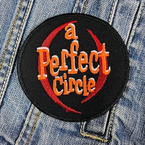 Puede incluir: Un parche circular negro con el texto "a Perfect Circle" en letras naranjas y blancas, rodeado por un diseño de media luna roja. El parche está sobre un fondo de mezclilla.
