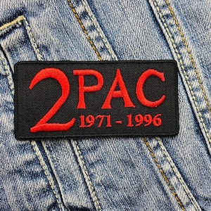 Puede incluir: Un parche rectangular negro con el texto rojo "2PAC" y los años "1971-1996". El parche está bordado sobre un fondo de mezclilla azul claro.