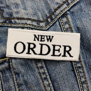 以下が含まれることがあります： 白い長方形のパッチに黒で「NEW ORDER」と刺繍され、デニムジャケットに付けられています。パッチは中央に配置され、テキストは太字のサンセリフフォントです。デニムジャケットはライトブルーウォッシュです。
