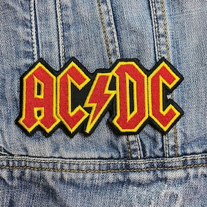 May include: Embroidered AC/DC logo patch in red and yellow, with a black border and lightning bolt graphic. The patch is in a bold, blocky font. It is sewn onto a denim jacket, with visible stitching and a blue wash.
