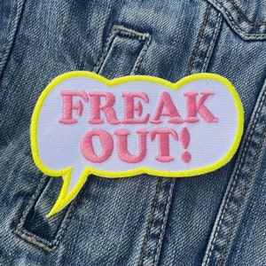 Könnte beinhalten: Gestickter Aufnäher in Sprechblasenform mit den Worten "FREAK OUT!" in rosa Schrift. Der Aufnäher hat einen weißen Hintergrund mit gelbem Rand und ist auf einer Jeansjacke befestigt.