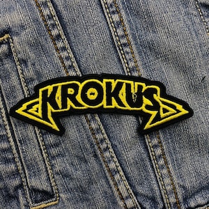 Könnte beinhalten: Schwarz-gelber gestickter Aufnäher mit dem Wort "KROKUS" in einem stilisierten Schriftzug. Der Aufnäher hat einen schwarzen Hintergrund mit gelben Buchstaben und pfeilförmigen Akzenten. Der Aufnäher befindet sich auf einer Jeansjacke.