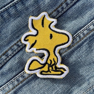 Può includere: Una toppa ricamata gialla raffigurante il personaggio dei cartoni animati Woodstock dei Peanuts. La toppa ha un bordo bianco e dettagli neri, su uno sfondo di denim blu.
