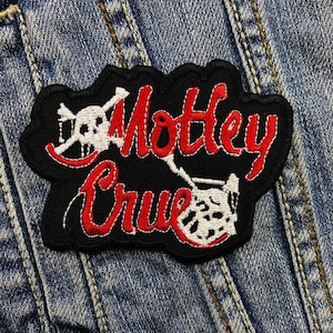 May include: A black patch with the words "Motley Crue" in red script. The patch features a white skull and crossbones design and a white drum set graphic. The patch is sewn onto a blue denim jacket.