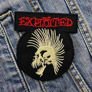 Pode incluir: Um patch preto com o texto "THE EXPLOITED" em vermelho acima de um patch redondo com uma caveira com moicano. A caveira e o moicano são de cor creme. Os patches são costurados em uma jaqueta jeans.