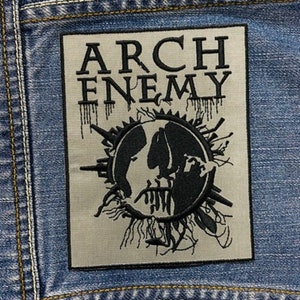 Peut inclure: Un écusson rectangulaire avec les mots "ARCH ENEMY" en noir, au-dessus d'un graphique noir et blanc d'un visage dans un cercle. L'écusson est cousu sur une veste en jean bleue.