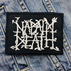 Puede incluir: Parche rectangular negro con texto bordado blanco "NAPALM DEATH" en una fuente gótica estilizada. El parche está sobre un fondo de mezclilla azul.