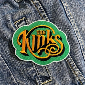 May include: Embroidered patch featuring the band name "The Kinks" in a stylized, gold-colored font with black accents. The patch has a green cloud-shaped background with a white border, set against a denim jacket.