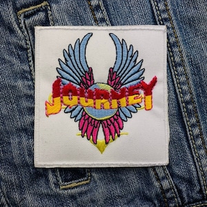 Könnte beinhalten: Gestickter Aufnäher mit dem Wort "JOURNEY" in Rot und Gelb, mit einem blauen und rosa Flügeldesign. Der quadratische Aufnäher ist auf eine Jeansjacke genäht. Der Aufnäher hat einen weißen Hintergrund und einen schwarzen Rand.
