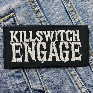 May include: A black rectangular patch with the words "KILLSWITCH ENGAGE" embroidered in white, gothic-style lettering. The patch is set against a denim background, showcasing the texture of the fabric and stitching.