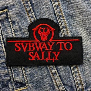 Könnte beinhalten: Schwarzer rechteckiger Aufnäher mit dem Text "SUBWAY TO SALLY" in Rot gestickt. Der Aufnäher zeigt ein rotes Bullenschädel-Design über dem Text. Der Aufnäher ist vor einem blauen Jeans-Hintergrund platziert.