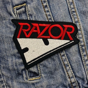 Pode incluir: Patch bordado com a palavra "RAZOR" em letras vermelhas sobre um desenho de lâmina de barbear branca. O patch tem uma borda preta e é costurado em uma jaqueta jeans.