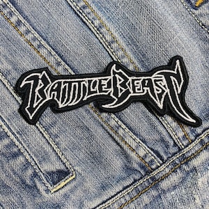 May include: Black and white embroidered patch with the text "BATTLE BEAST" in a stylized font. The patch is rectangular with a pointed design on each end. The patch is on a denim background.