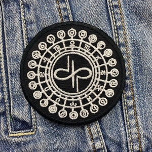 Puede incluir: Un parche redondo negro con un diseño circular blanco. El diseño presenta un símbolo central dentro de un círculo, rodeado por un anillo exterior de símbolos y formas. El parche está sobre un fondo de mezclilla azul.