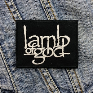 May include: A black rectangular patch with the white text "lamb of god". The patch is on a blue denim jacket. The text is in a stylized font.