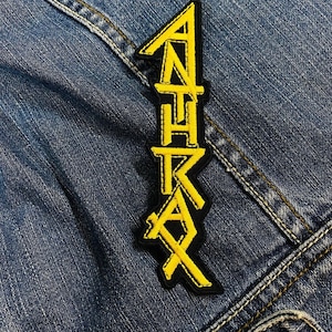 May include: A black and yellow embroidered patch with the word "ANTHRAX" in a vertical design. The patch is on a denim jacket.
