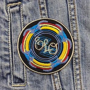 Puede incluir: Parche bordado circular con el logotipo de la banda "ELO", en un diseño estilizado con detalles azules, amarillos y rojos. El parche está adherido a una chaqueta vaquera, lo que demuestra su uso para la personalización de ropa.