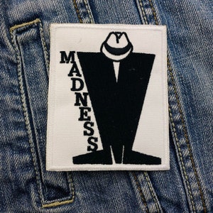 Könnte beinhalten: Ein quadratischer, weißer Aufnäher mit dem Wort "MADNESS" in schwarzen, vertikalen Buchstaben. Eine schwarze Figur in Anzug und Hut ist auf dem Aufnäher zentriert. Der Aufnäher befindet sich auf einer Jeansjacke.