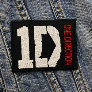 May include: A black rectangular patch featuring the letters "1D" in white and the words "ONE DIRECTION" in red. The patch is set against a denim background.